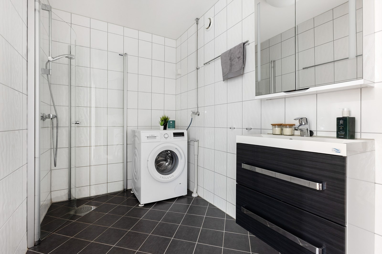Badet har fliser på både gulv og vegger. Innredningen inkluderer et servantskap i mørk, glatt utførelse med en heldekkende servant. Galleribilde