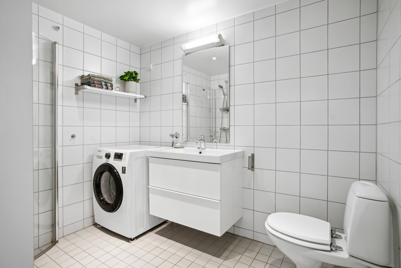Lyst, flislagt bad fra byggeår. Rommet er innredet med dusjhjørne med 2 stk svingbare glassdører, baderomsinnredning med slette fronter og vegghengt toalett. Galleribilde