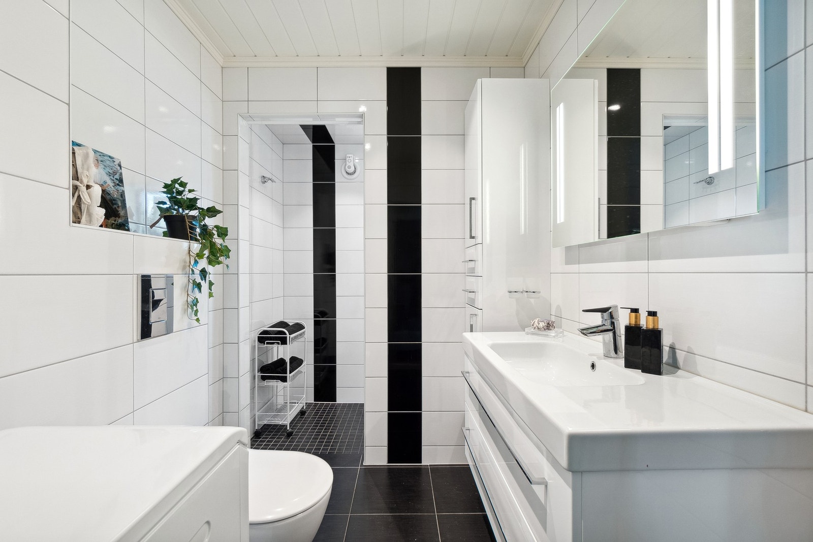 Badet har hvit innredning med god plass for oppbevaring, Heldekkende servant, veggmontert toalett, dusjnisje og opplegg for vaskemaskin. Galleribilde