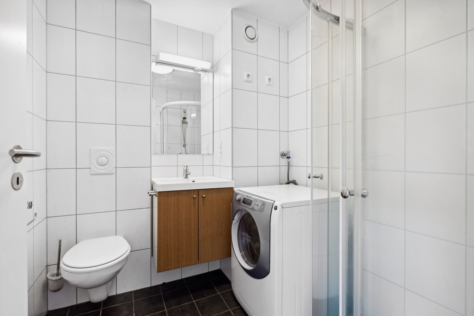 Flislagt baderom med vegghengt toalett, servant, dusj og opplegg til vaskemaskin Galleribilde