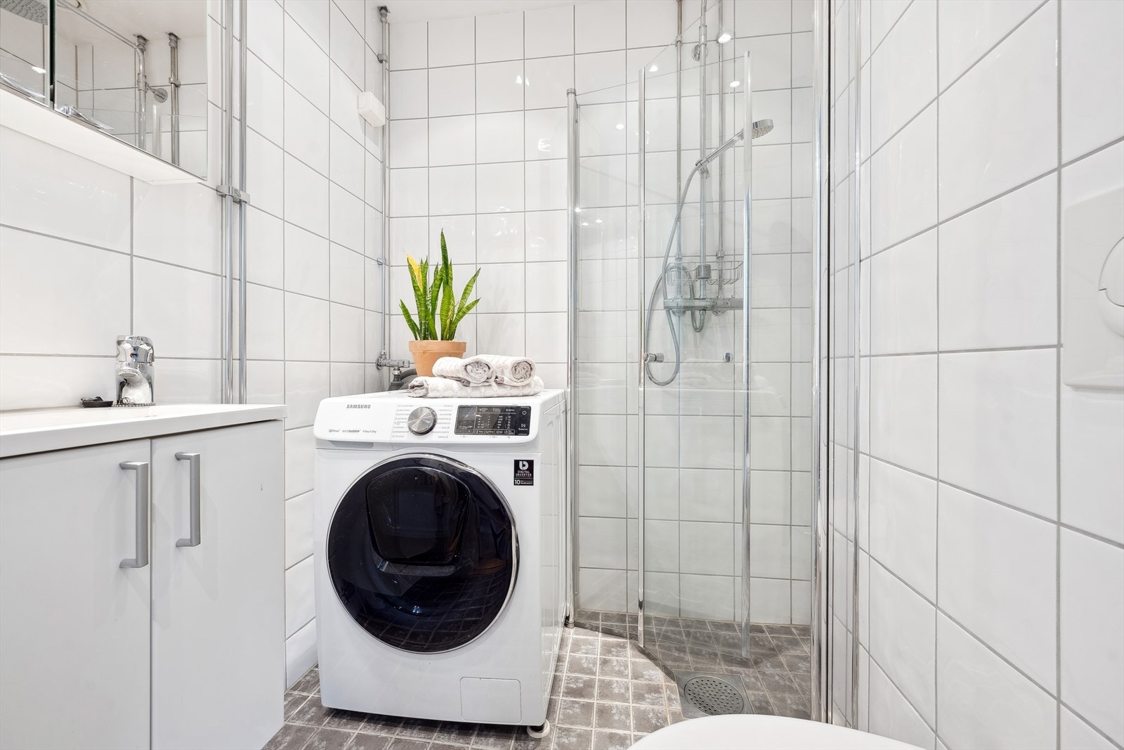 Flislagt baderom fra 2009 med servantskap heldekkende servant, speilskap med lysarmatur, veggmontert klosett, dusjhjørne og opplegg for vaskemaskin. Galleribilde