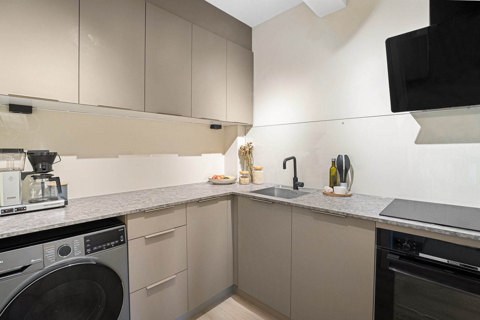 KVIK kjøkken med glatte fronter og benkeplate av laminat, nedsenket oppvaskkum med ett-greps armatur, malt flate og glass mellom benkeplate og overskap, stikkontakter under overskap og vegghengt ventilator med kullfilter. Galleribilde