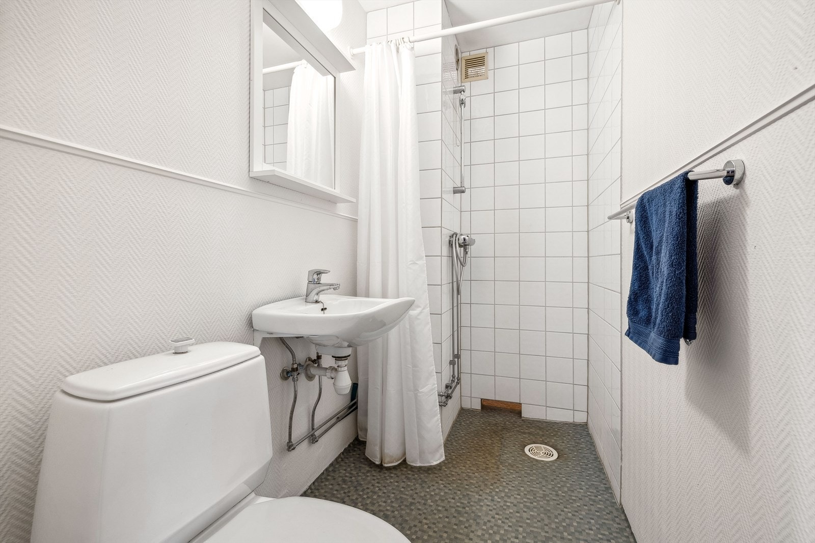 Eldre baderom med behov for full modernisering. Baderommet har flislagt våtsone, belegg på gulv og malt tapet ellers i rommet. Rommet består av servant, speil og lysarmatur montert påvegg, gulvmontert toalett og dusjhjørne med forheng. Galleribilde