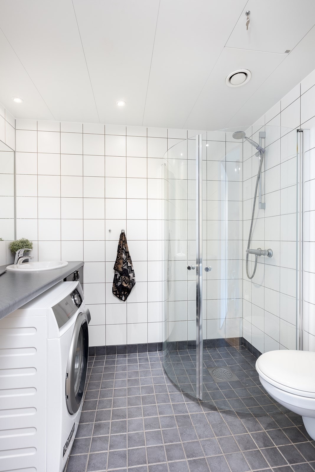 Badet består av vegghengt servantskap, benkeplate av laminat, nedfelt servant, innfelt speil, stikkontakt på vegg over servant, dusjsone med innfellbare dører av glass, dusjarmatur tilkoblet hånddusj og vegghengt toalett. Nedsenket himling med downlight Galleribilde