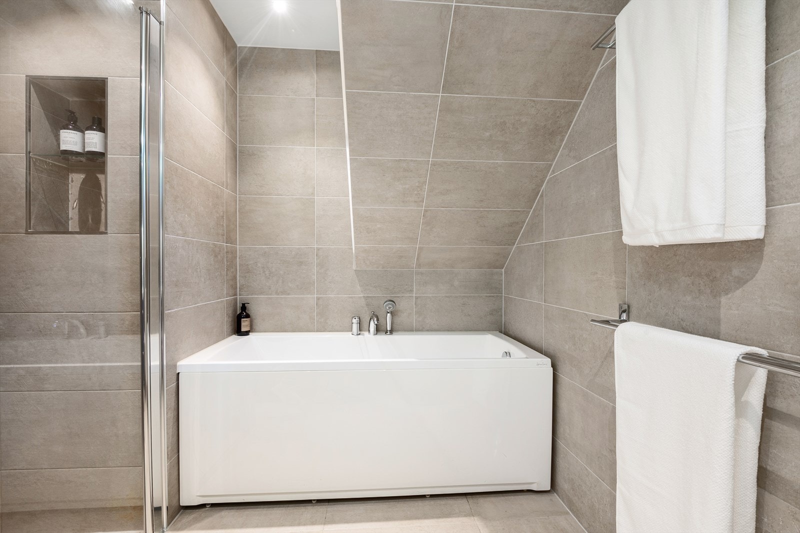 Rommet har badekar i tillegg til dusjnisje med glassdør, innebygd hylle, regnfallsdusj og mosaikkflis på gulv og vegg. Videre består innredningen av veggmontert toalett, bred nedfelt servant over skuffesekson og dobbelt speilskap med lys. Galleribilde