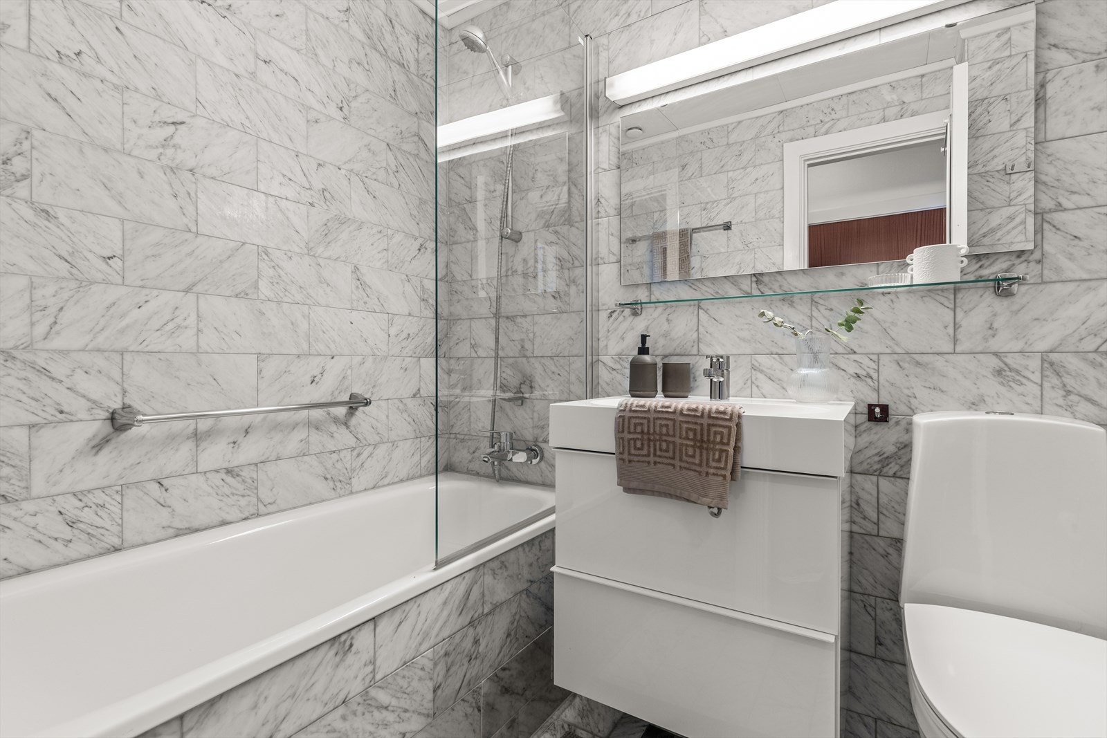 Delikat flislagt baderom med marmorfliser, badekar/dusjnisje, wc, servant-skap og speil. Galleribilde