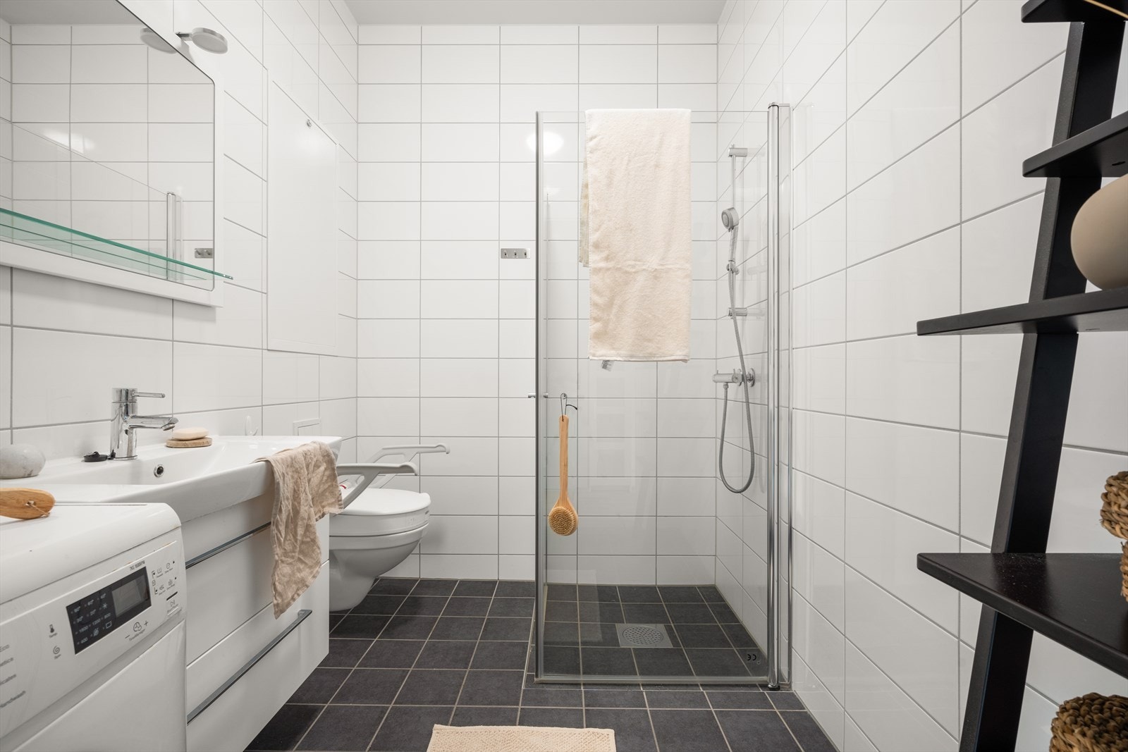 Badet er utstyrt med vegghengt toalett, helstøpt servant, skuffer med slette fronter under servant , dusjdører i klart glass, opplegg for vaskemaskin og fordelerskap til rørsystemet Galleribilde