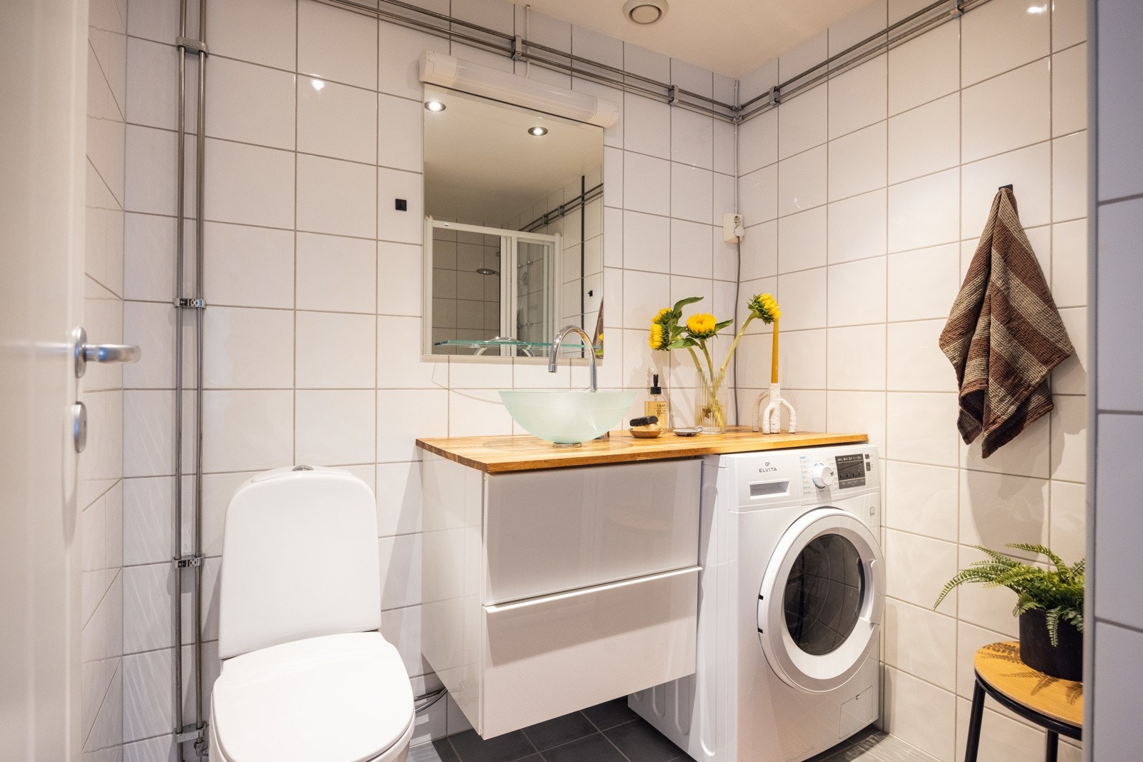 Fint, flislagt baderom som består av toalett, servant over innredning, speil og dusjhjørne. Galleribilde