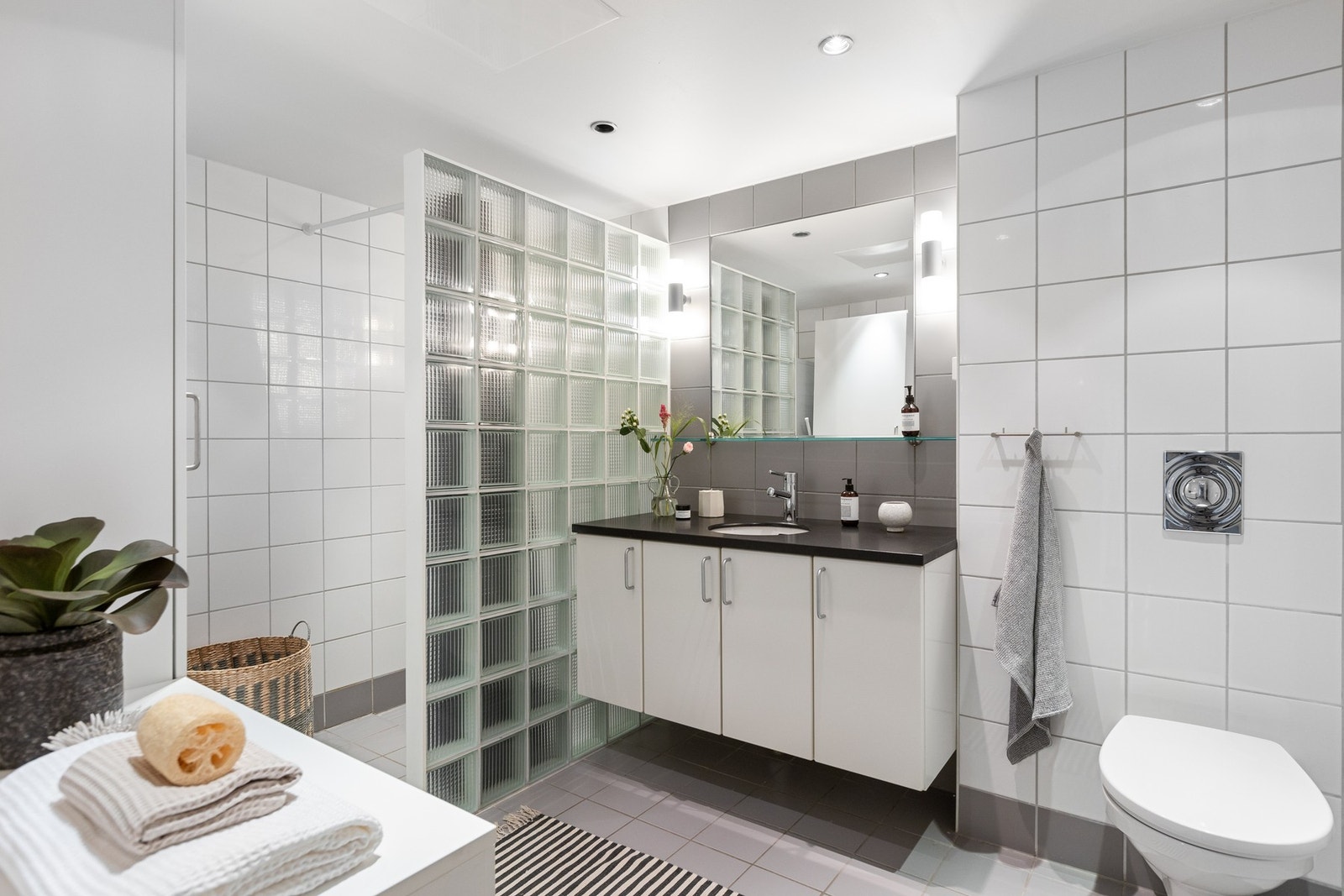 Romslig baderom for velvære har varme i gulvstøp med grå fliser på gulv og hvite fliser på vegg. Det er vegghengt toalett med innebygget sisterne for enkel rengjøring. Galleribilde