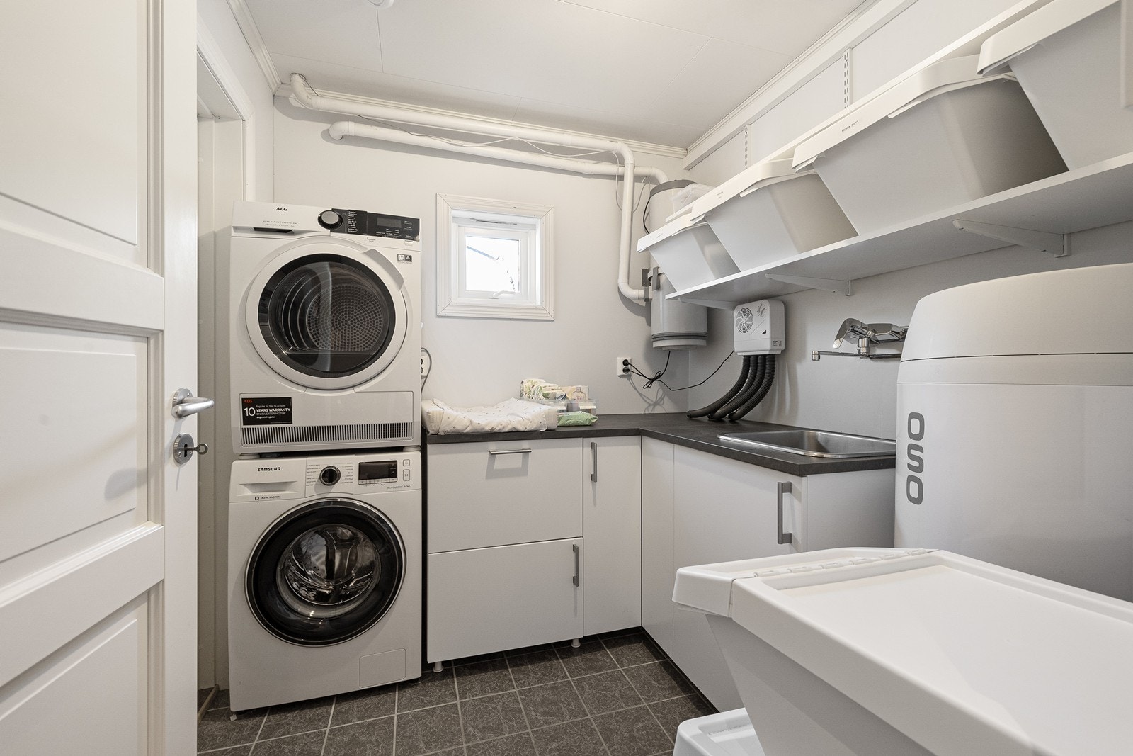 Praktisk vaskerom med benkeinnredning, hyller, varmtvannsbereder, opplegg for vaskemaskin og tørketrommel og sentralstøvsuger. Galleribilde