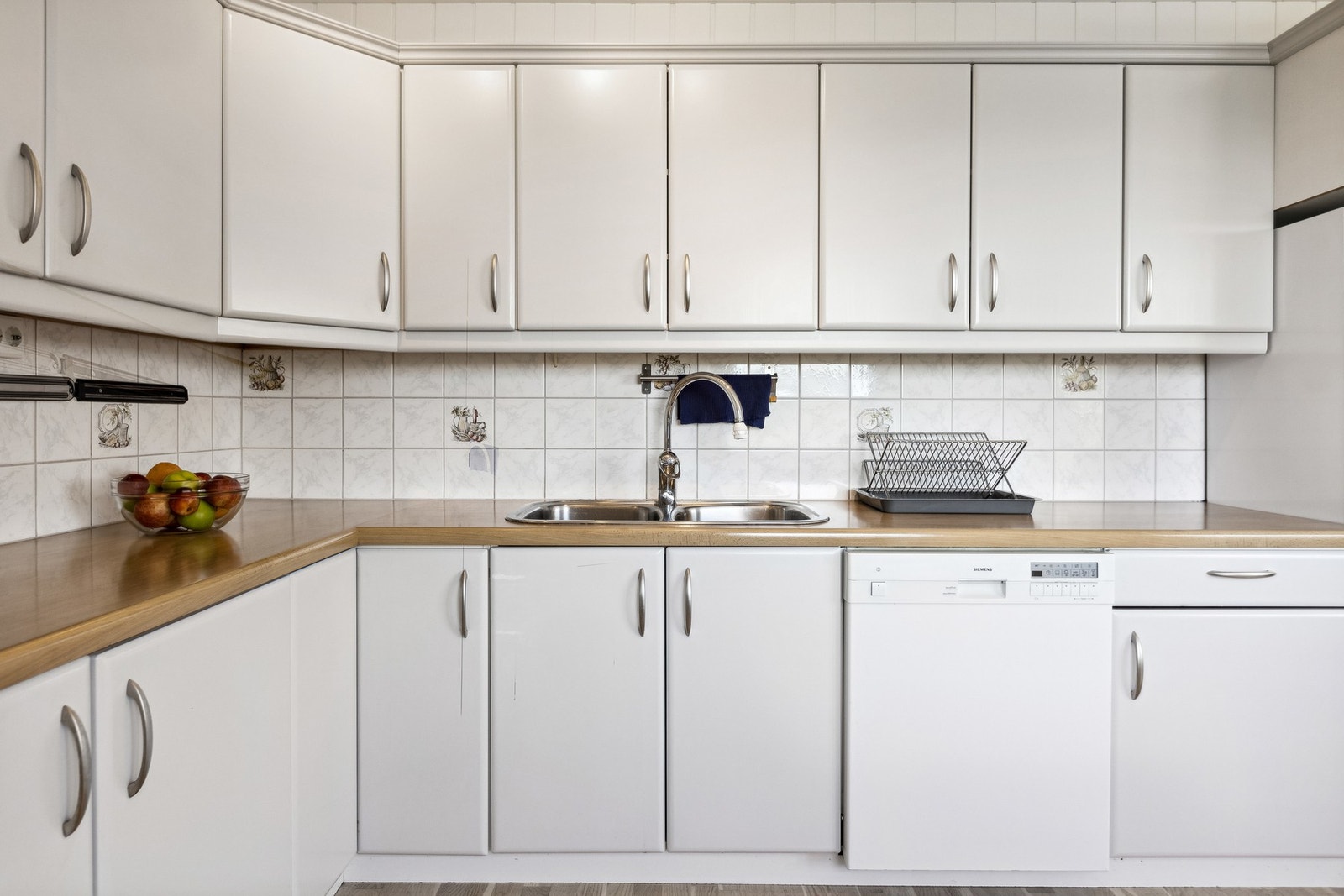 Hvite, slette fronter på skap/skuffer. Benkeplate av laminat med nedfelt vaskekum/beslag av stål og det er keramiske fliser over kjøkkenbenk Galleribilde