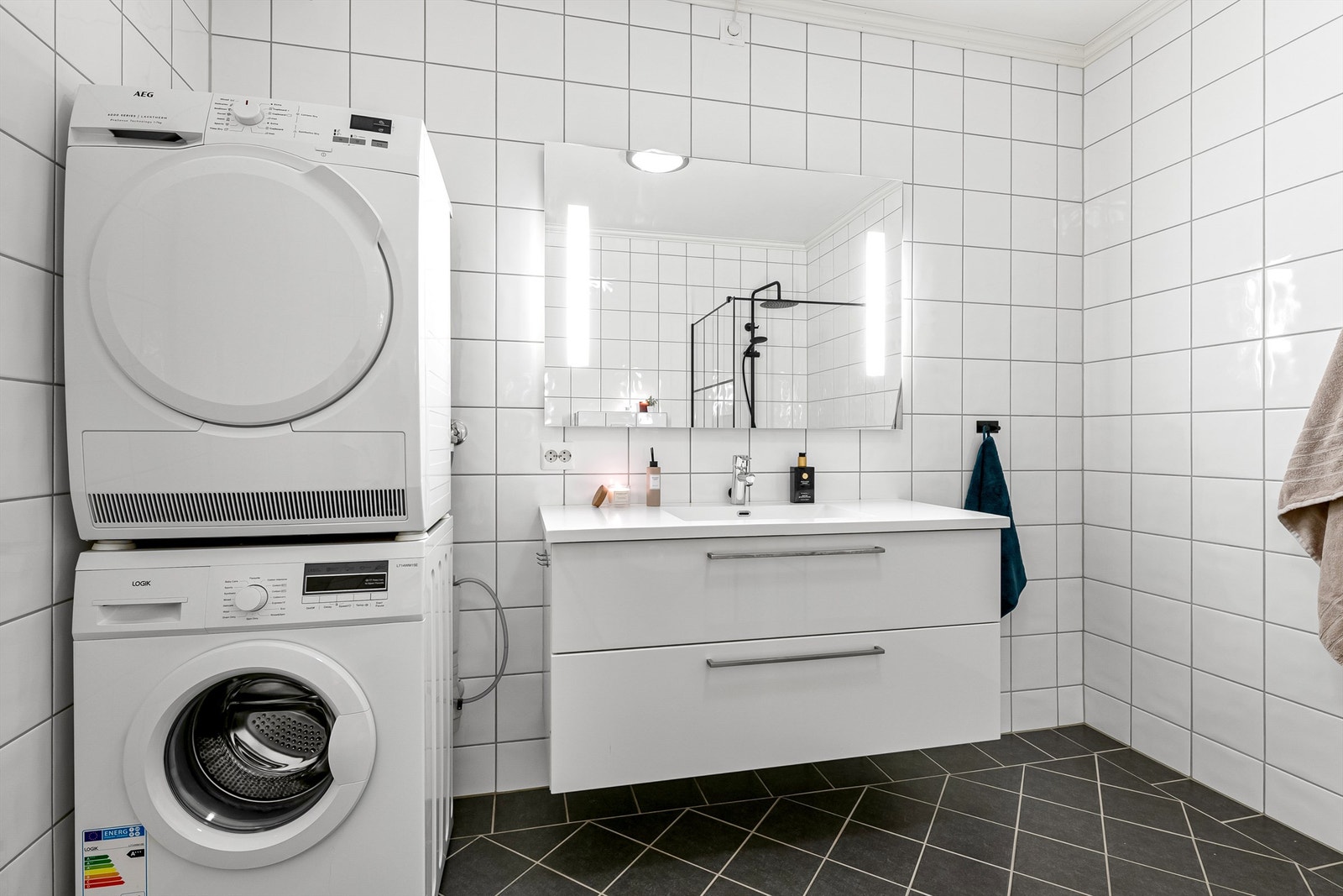 Hvit baderomsinnredning med skuffer og speil med lys over servant. Det er opplegg for vaskemaskin og tørketrommel på badet Galleribilde
