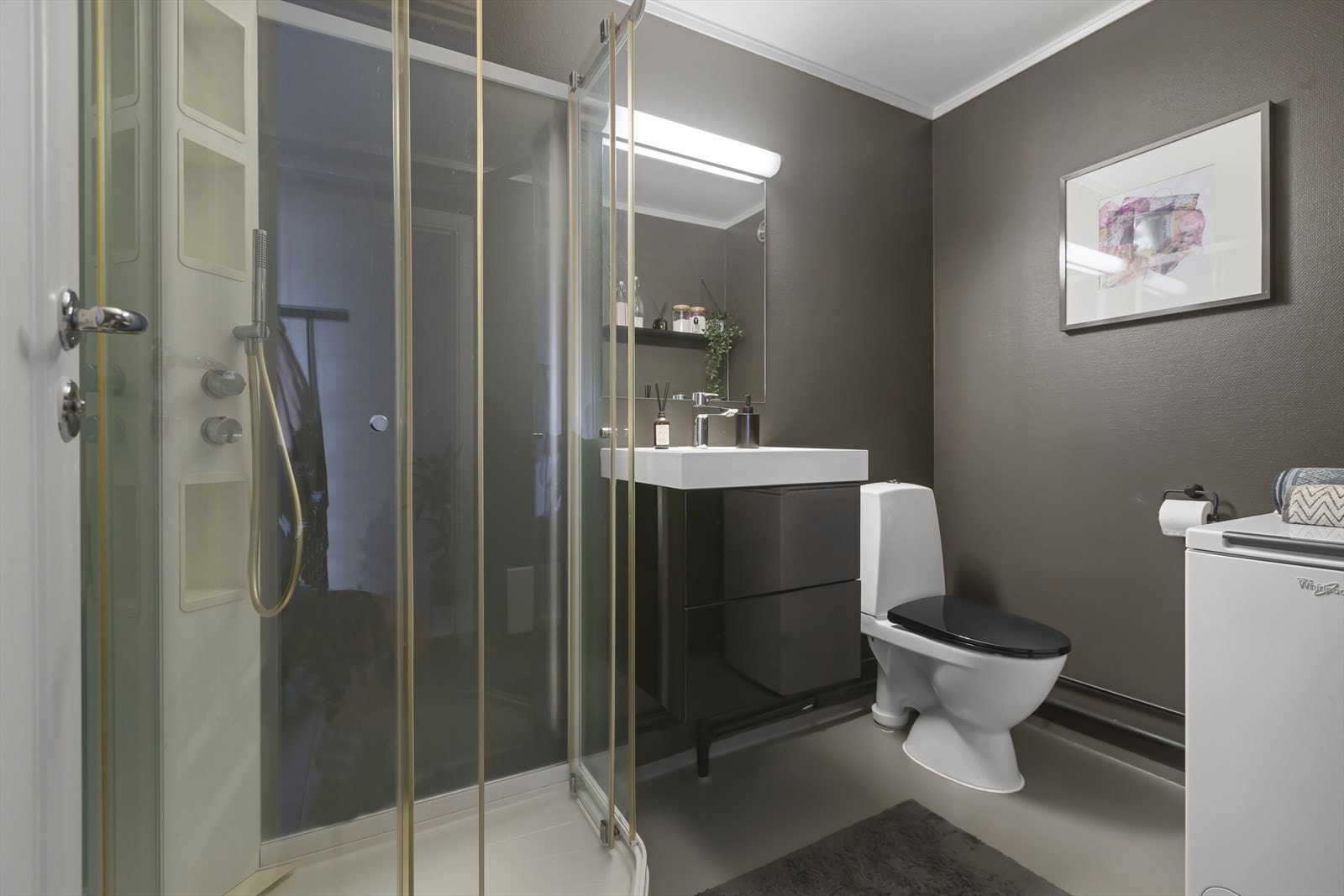 Badet har belegg på gulvet og mørkmalt glassfiberstrie på veggene. Rommet er praktisk utstyrt med toalett, servantinnredning og dusjkabinett. Galleribilde