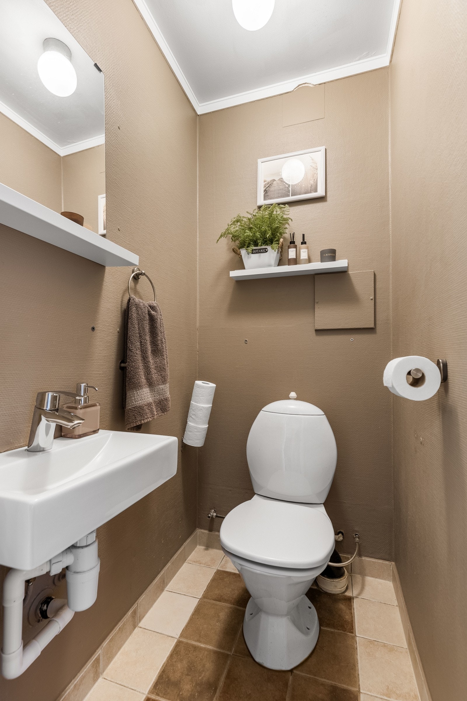 Vegg-i-vegg med badet i underetasjen ligger et pent toalettrom med hvite gulvfliser, sokkelflis, strietapet malt i en varm farge, gulvvarme og balansert avtrekk. Galleribilde