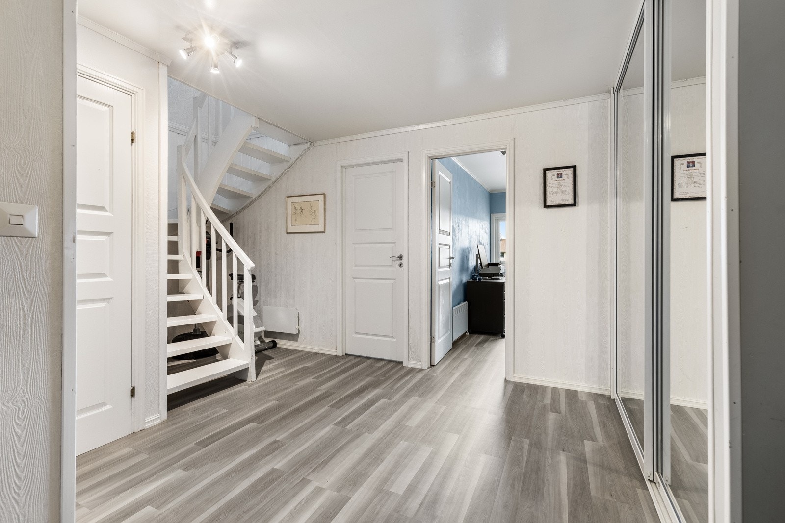 En åpen, hvitmalt tretrapp gir adkomst ned til den store gangen i etasjen under som har en romslig skyvedørsgarderobe med speilfronter. Galleribilde