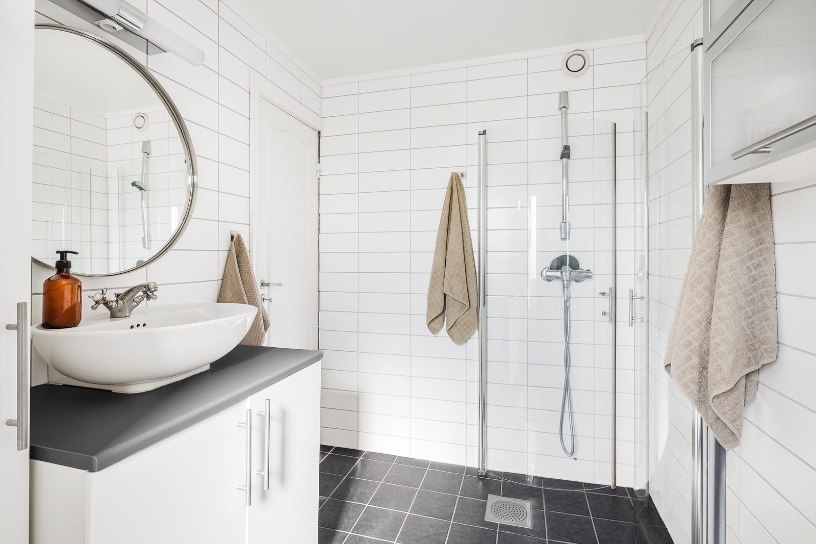Tidløs baderomsinnredning med hvite fronter og laminert benkeplate med ovenpåliggende servant, rundt speil med belysning over. Baderommet er også utstyrt med dusjhjørne med innfellbare glassdører. Galleribilde