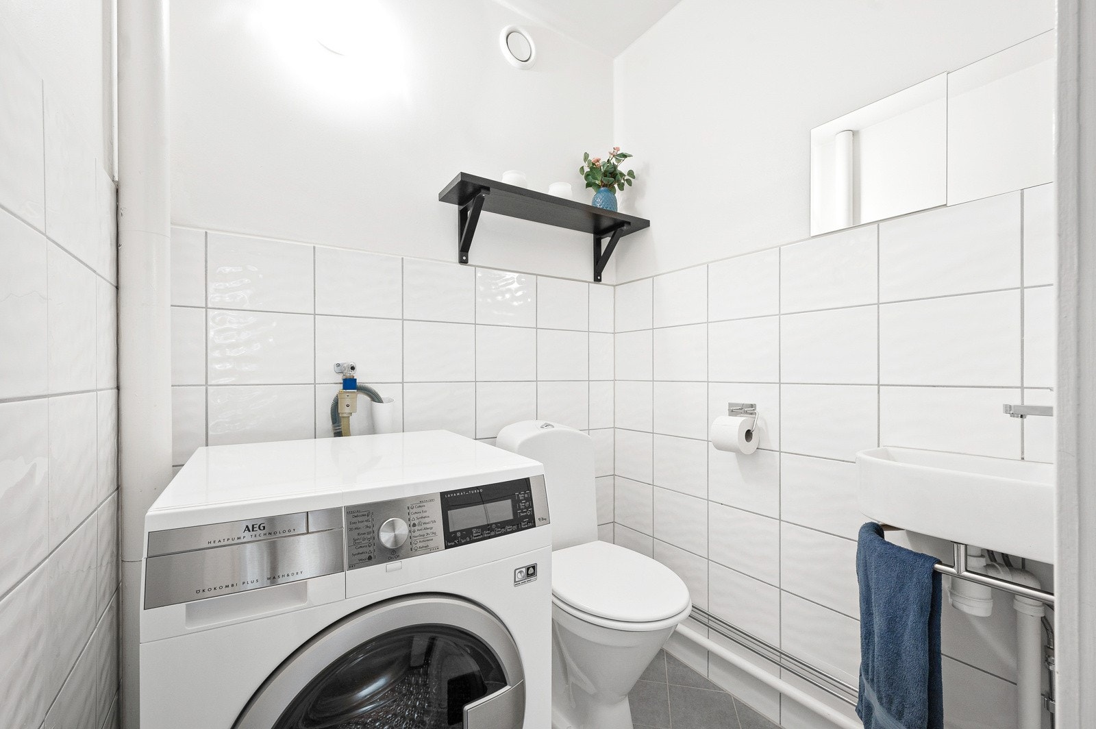 Toalettrom/vaskerom med gulvstående toalett, vegghengt servant og opplegg for vaskemaskin. Galleribilde
