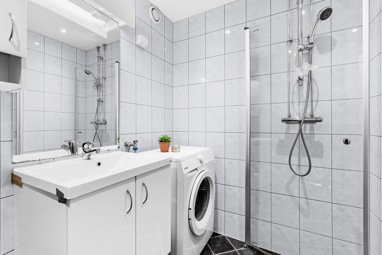 Badet har også opplegg for vaskemaskin, noe som bidrar til praktisk håndtering av klesvasken. Dusjhjørnet med rette glassdører gir en moderne følelse og er plassert i en egen sone med sluk. Galleribilde
