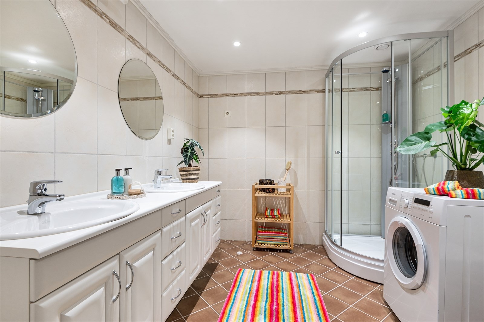 Badet er flislagt fra topp til tå, det er utstyrt med dusjkabinett, opplegg for vaskemaskin, rørfordelingsskap, servant, servantskap og veggmontert wc. Galleribilde