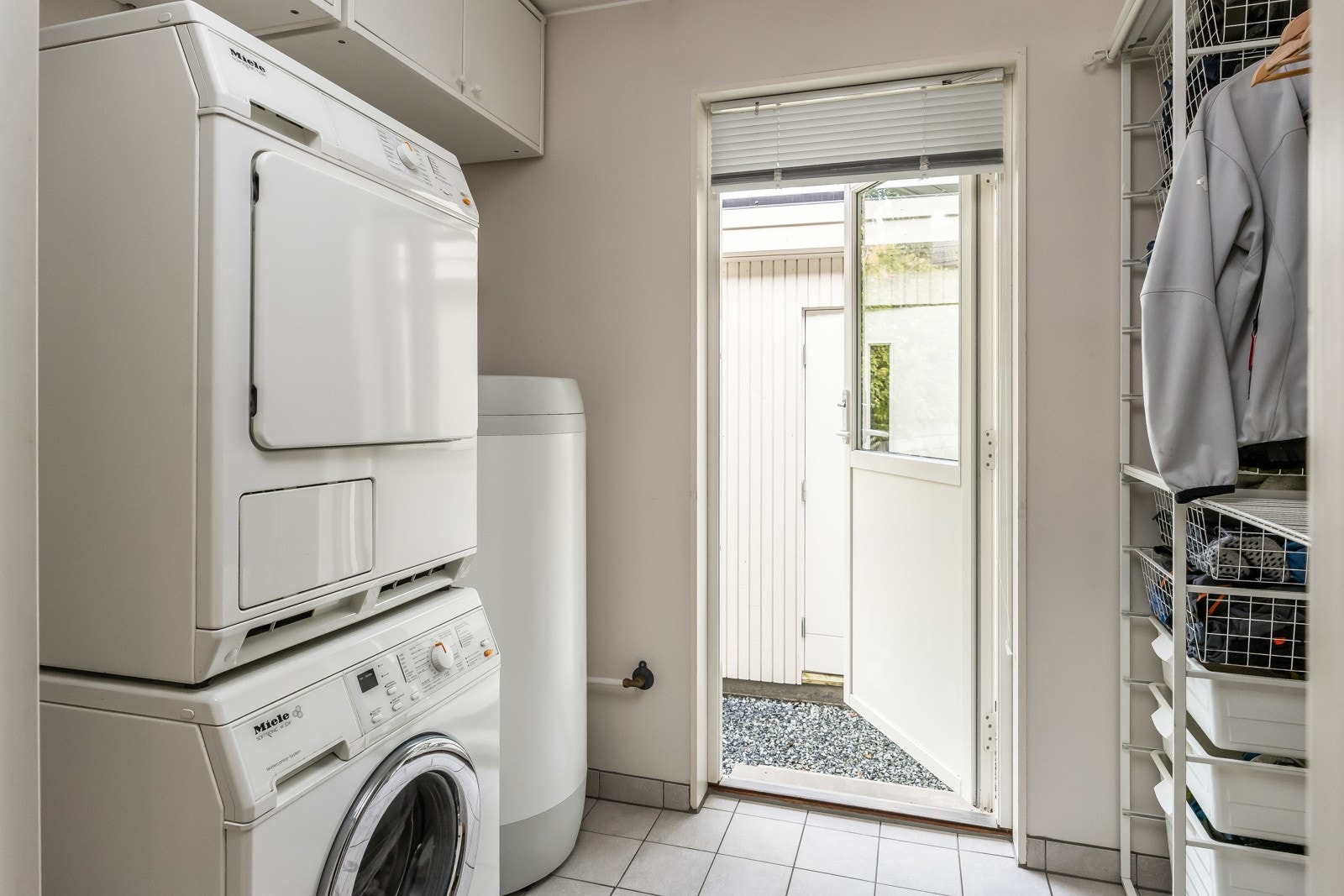 Praktisk vaskerom med egen utvendig inngang. Rommet har fliser på gulv og har opplegg for vaskemaskin/tørketrommel, hyllesystem og varmtvannsbereder. Galleribilde