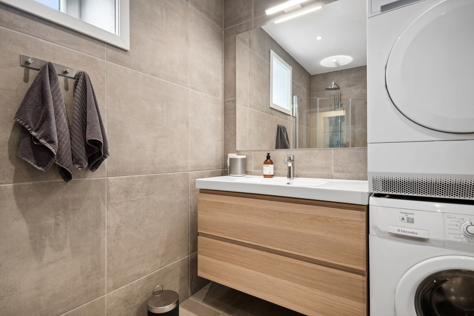 Badet inneholder dusjhjørne med glassdører, vegghengt toalett, servant med skuffer, speil på vegg og opplegg for vaskemaskin og tørketrommel. Galleribilde