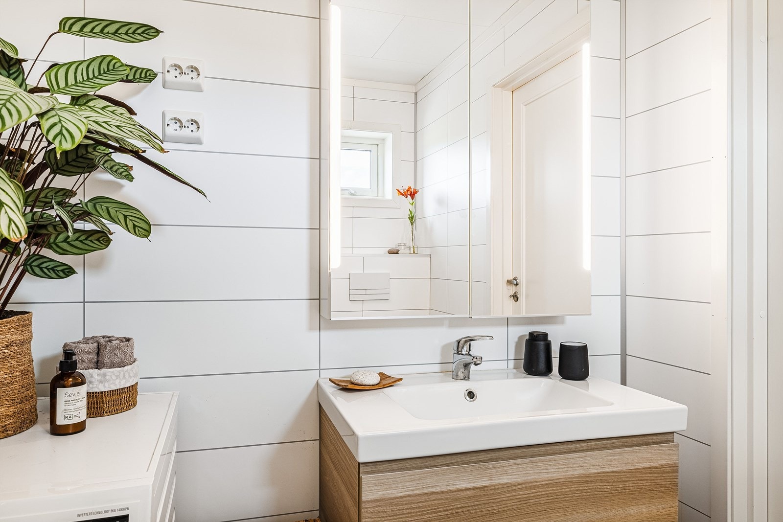 Moderne baderomsinnredning med skuffer og heldekkende servant, vegghengt speilskap med integrert belysning over. Plass og opplegg for vaskemaskin. Galleribilde