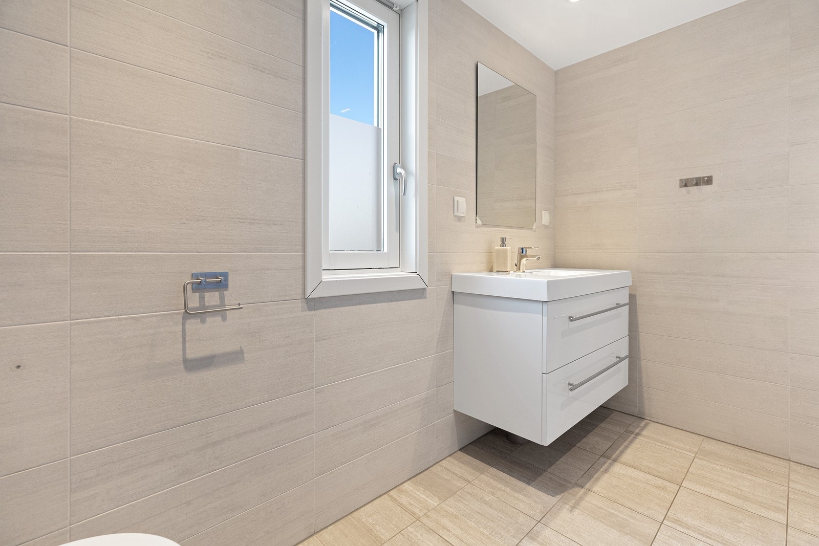 Badet er lyst og delikat flislagt bad med veggmontert toalett, servant med baderomsinnredning og dusjhjørne med innadslående glassdører. Galleribilde
