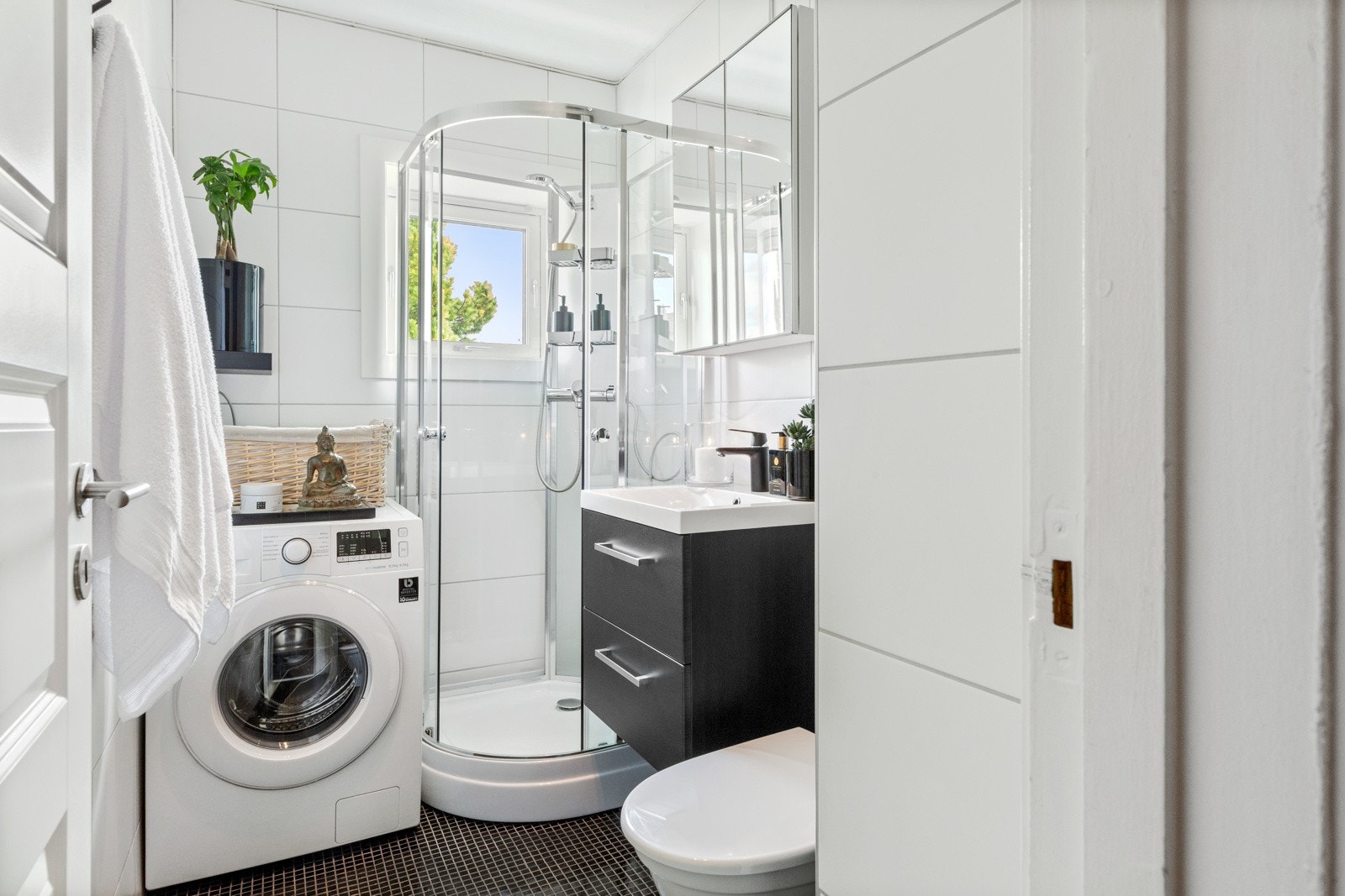 Lyst baderom med dusjkabinett, opplegg til vaskemaskin og toalett. Badet har en moderne innredning. Galleribilde