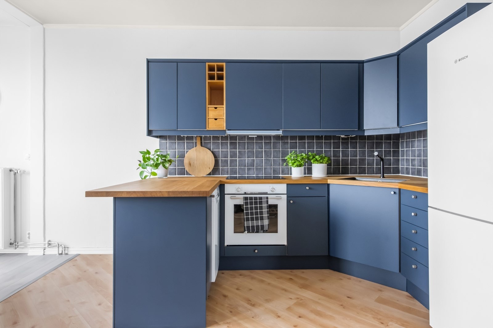 Kjøkkeninnredning med glatte fronter og benkeplate. Flislagt vegg over benkeskap. Kjøkkenet er utstyrt med platetopp, kum med ettgreps blandebatteri, ventilator, kjøl/fryseskap, ovn og oppvaskmaskin. Galleribilde