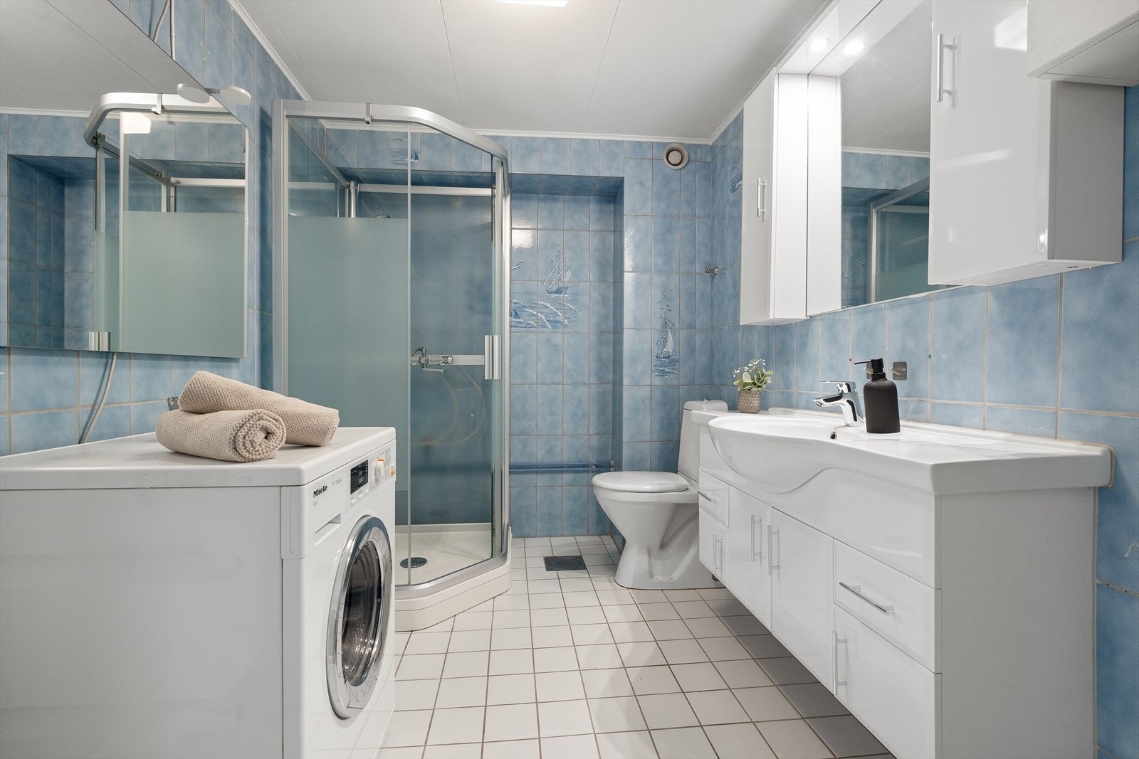 Pent og stilrent bad med fliselagt gulv og varmekabler. Det er stående fliser, kombinert med malte innvendige plater på vegger. Badet har wc, dusjkabinett, servant i innredning med overskap samt speil og lys i rommet. Opplegg til vaskemaskin på badet. Galleribilde