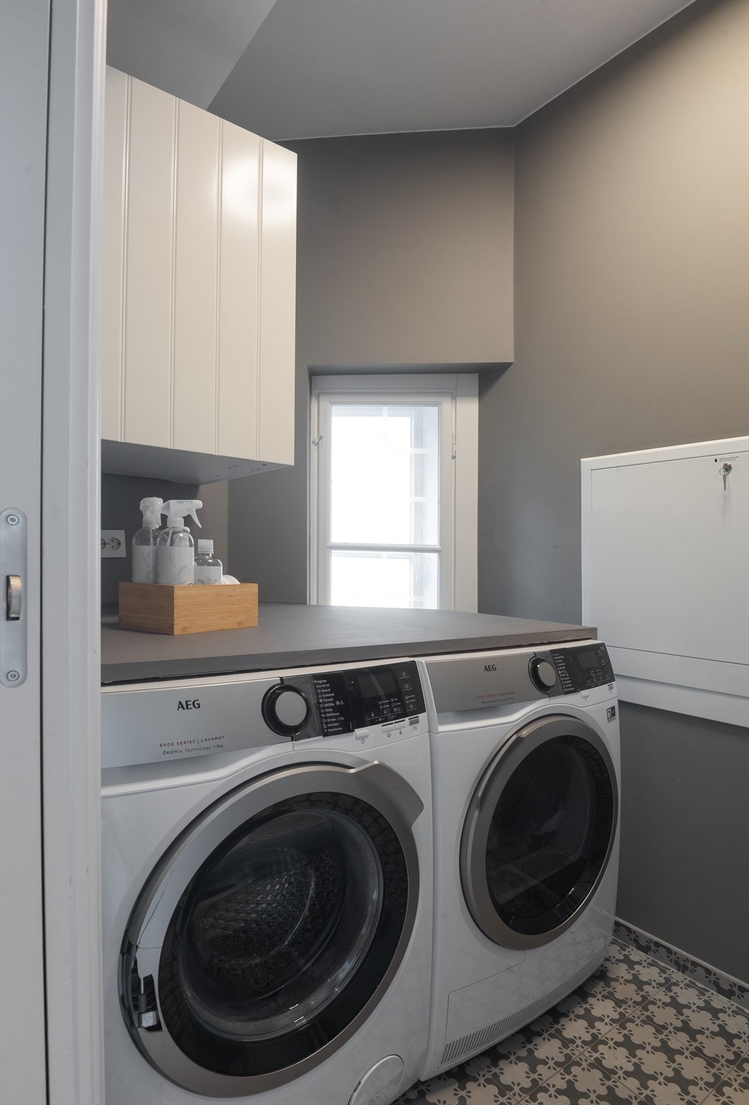 Teknisk rom/vaskerom med opplegg for vaskemaskin/tørketrommel, flislagt gulv med sluk, el. vifte montert i himling, benkeplate og veggskap. Galleribilde