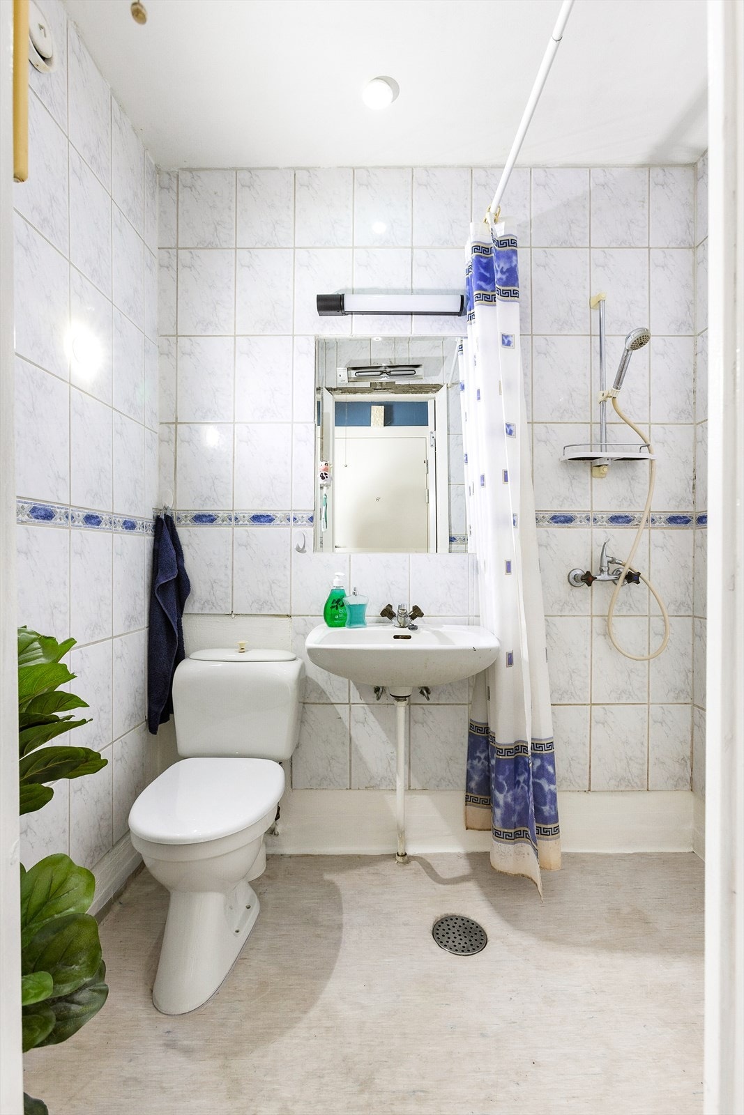 Eldre bad med belegg på gulv og fliser på vegger. Badet er utstyrt med servant, speil med belysning over, gulvstående toalett og dusjhjørne med veggmontert armatur og garnityr. Galleribilde