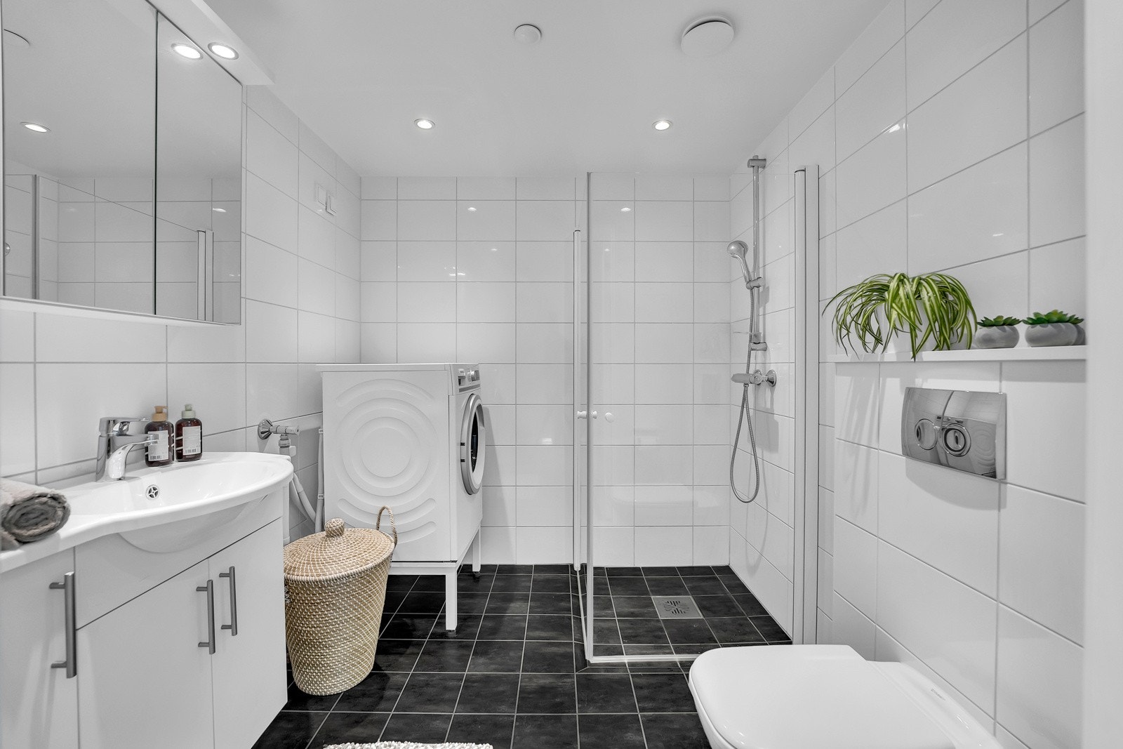 Badet har sorte fliser på gulvet og hvite delikate fliser på veggene. Videre kan badet skilte med gulvvarme, spotter i himlingen, opplegg for vaskemaksin, plass til tørketrommel, vegghengt toalett, dusj med innfellbare dører og servant. Galleribilde