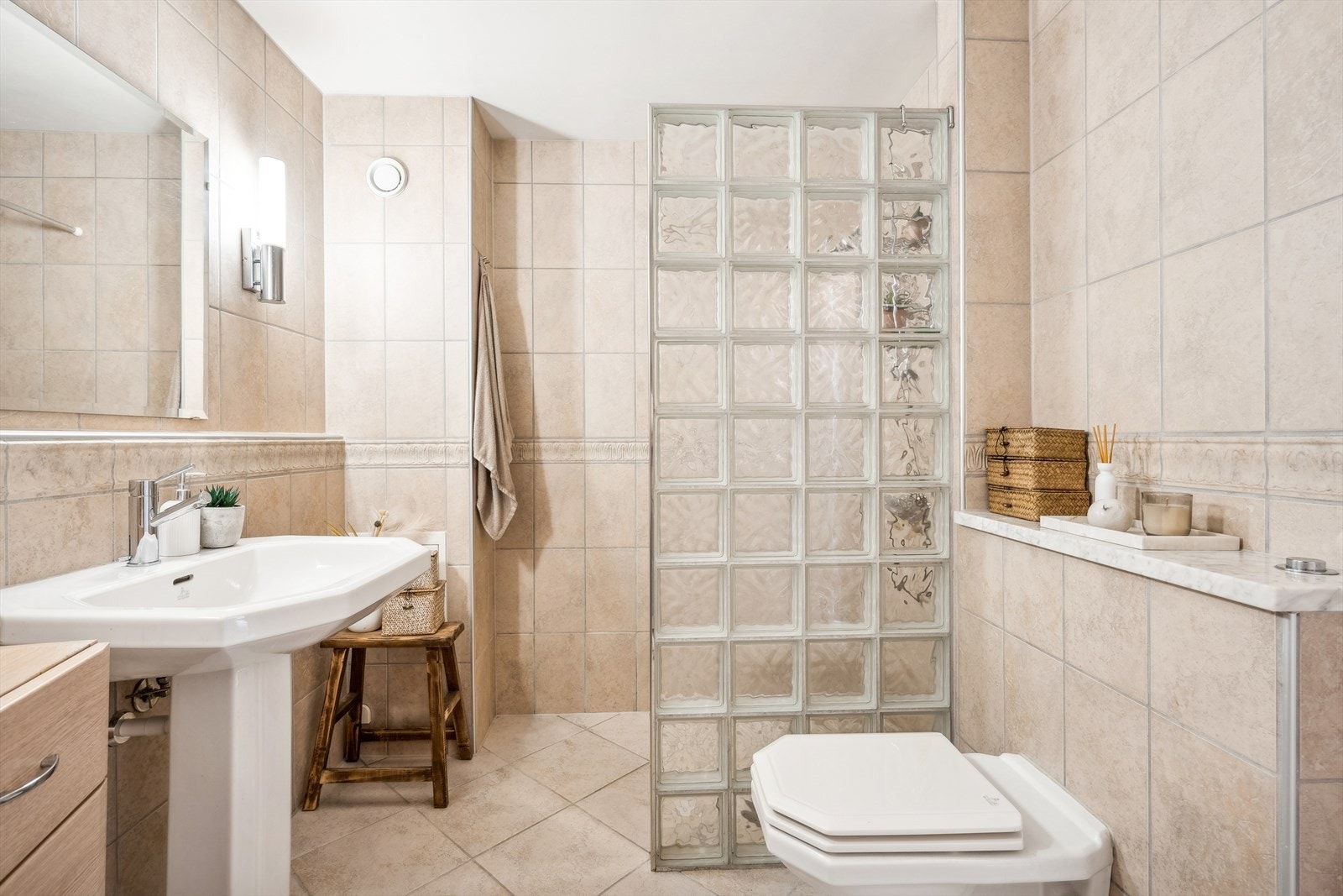Badet er innredet med vegghengt servant med søyle, ett-greps blandebatteri, vegghengt toalett med innebygget sisterne, avskjermet dusjhjørne med glassbyggerstein, blandebatteri og dusjgarnityr. Galleribilde