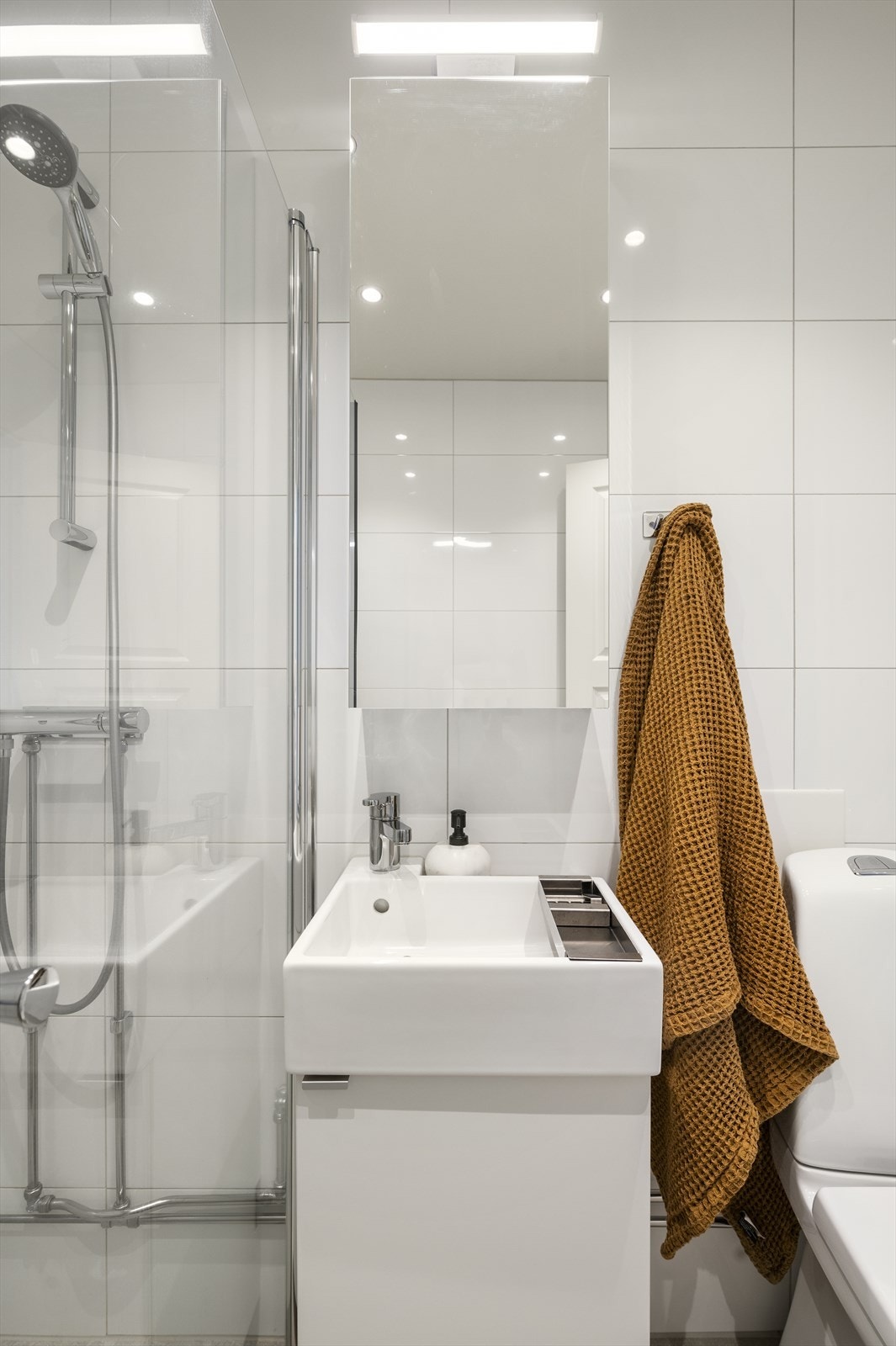 Badet er utstyrt med et vegghengt klosett, en servant nedfelt i innredningen, speilskap med belysning og et dusjhjørne med svingbare dører Galleribilde