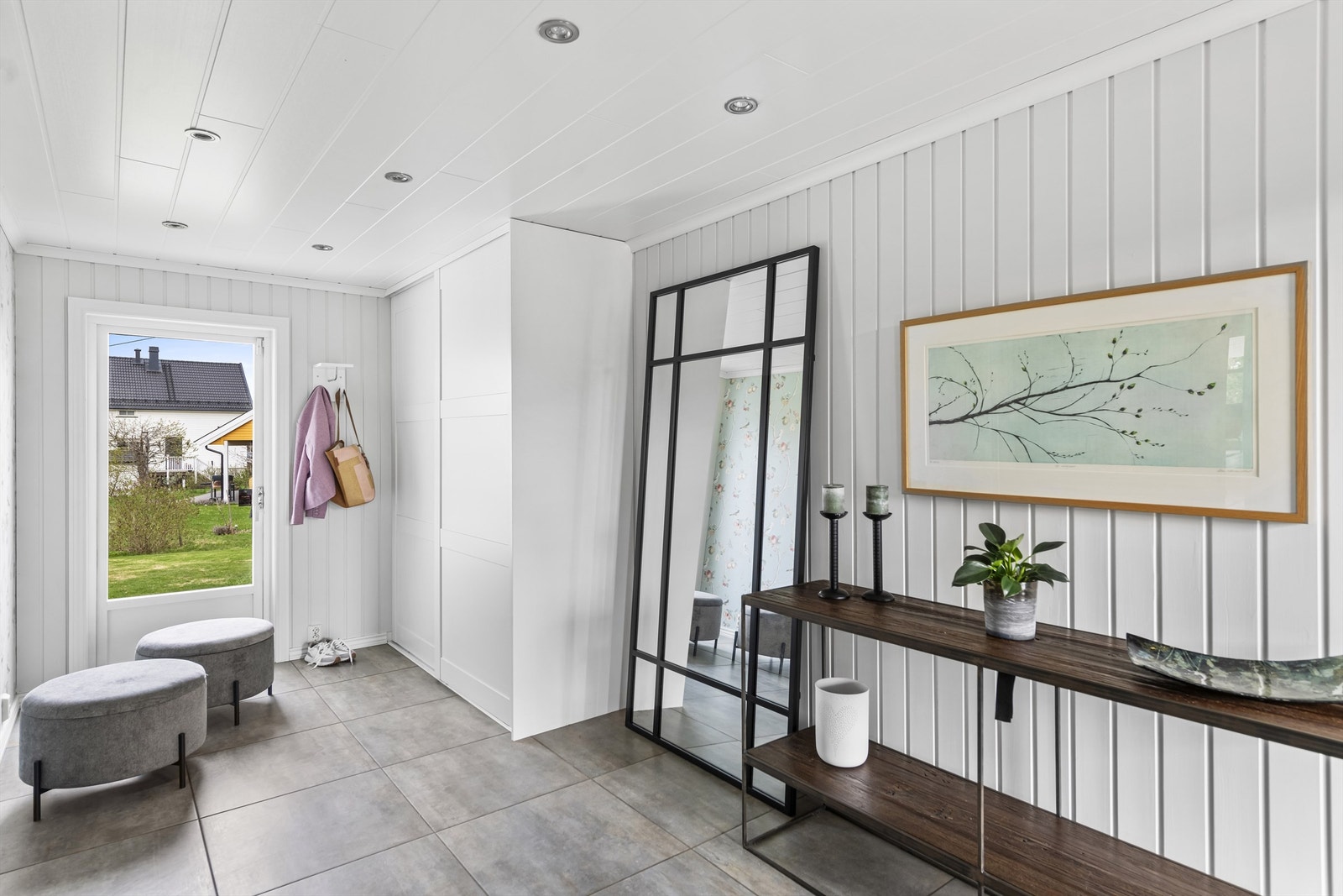 Entreen har mørke fliser på gulv, hvitmalte panelplater med downlights i himling, lysmalte panelvegger, og en kledelig fondvegg med mønstertapet i en sart grønntone. En takhøy skyvedørsgarderobe tar seg av yttertøy og sko. Galleribilde