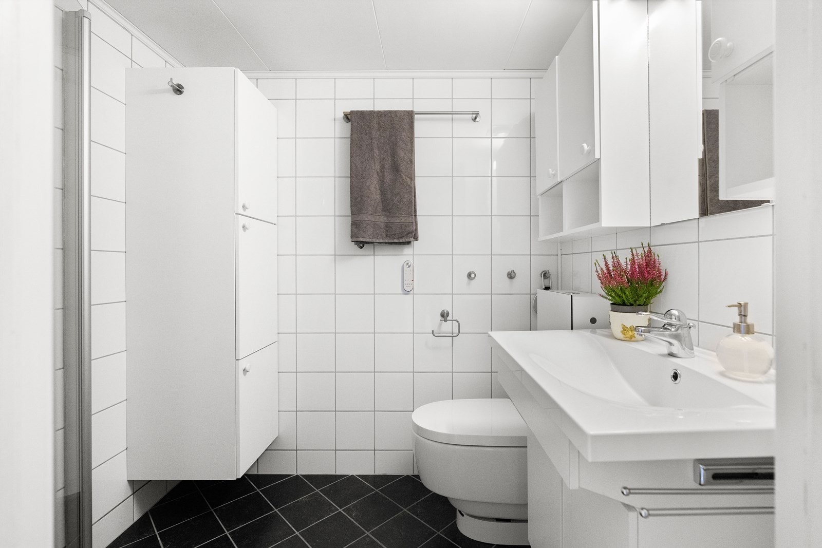 Videre er rommet utstyrt med dusj på gulv med dører i herdet glass, veggfestet klosett med veggmontert sisterne samt servant med benkeskap Det er ekstra lagringsplass i veggskap med glatte fronter. Galleribilde