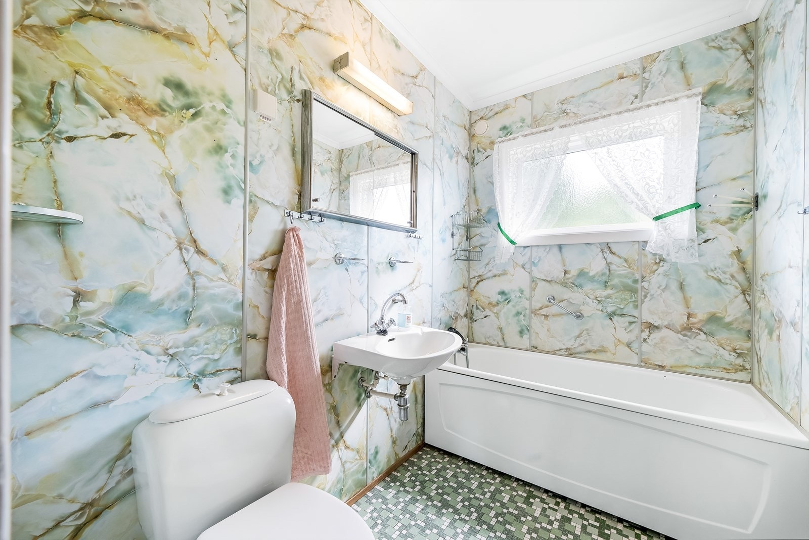 Eldre bad med baderomsplater på vegg og fliser på gulv. Innredet med servant med speilskap, toalett og badekar med hånddusj. Galleribilde