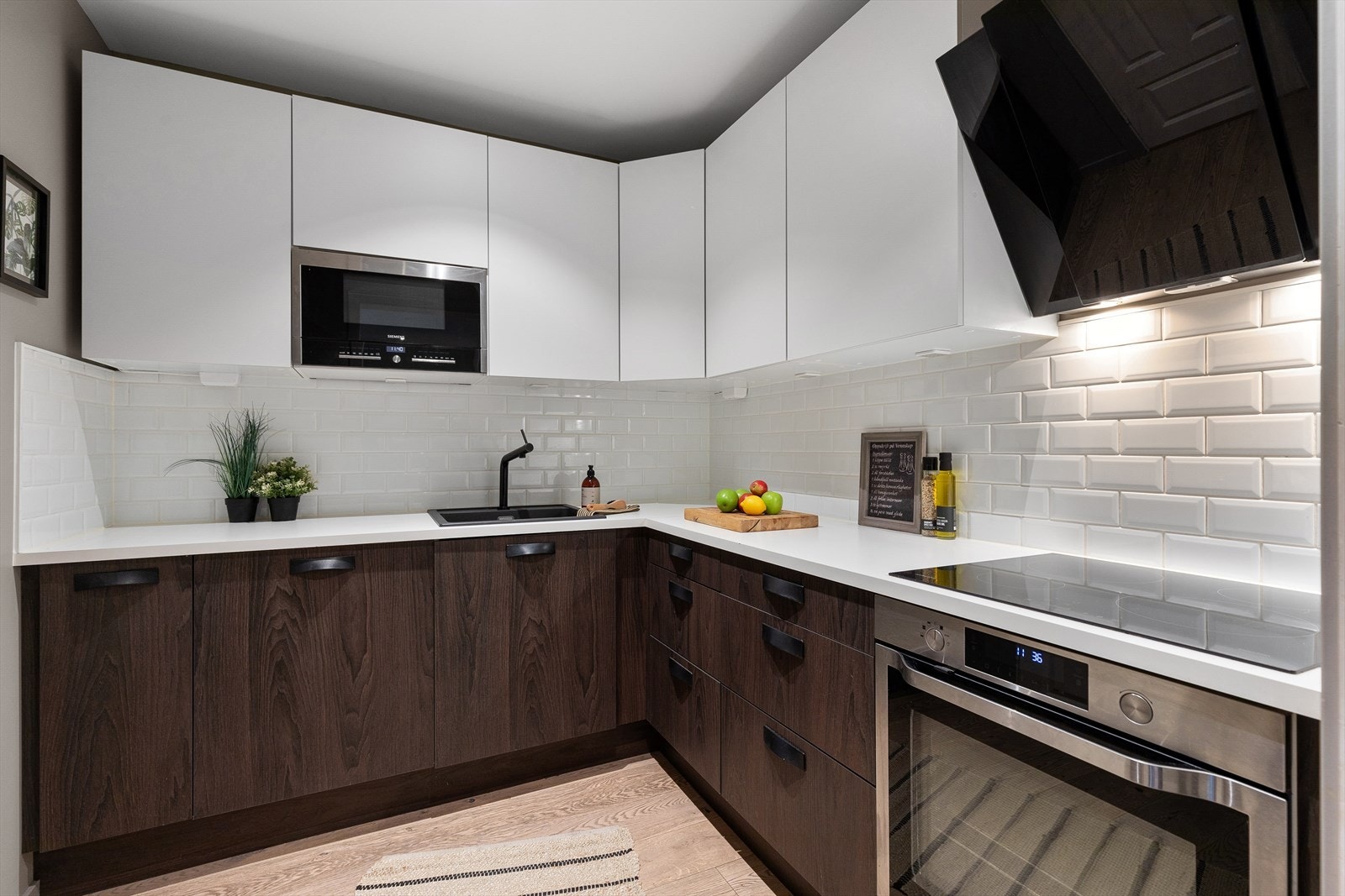 Kjøkken med store overskap med hvite glatte fronter og romslige benkeskap med mørke laminerte fronter i imitert tre. Hvite laminerte benkeplater. Galleribilde