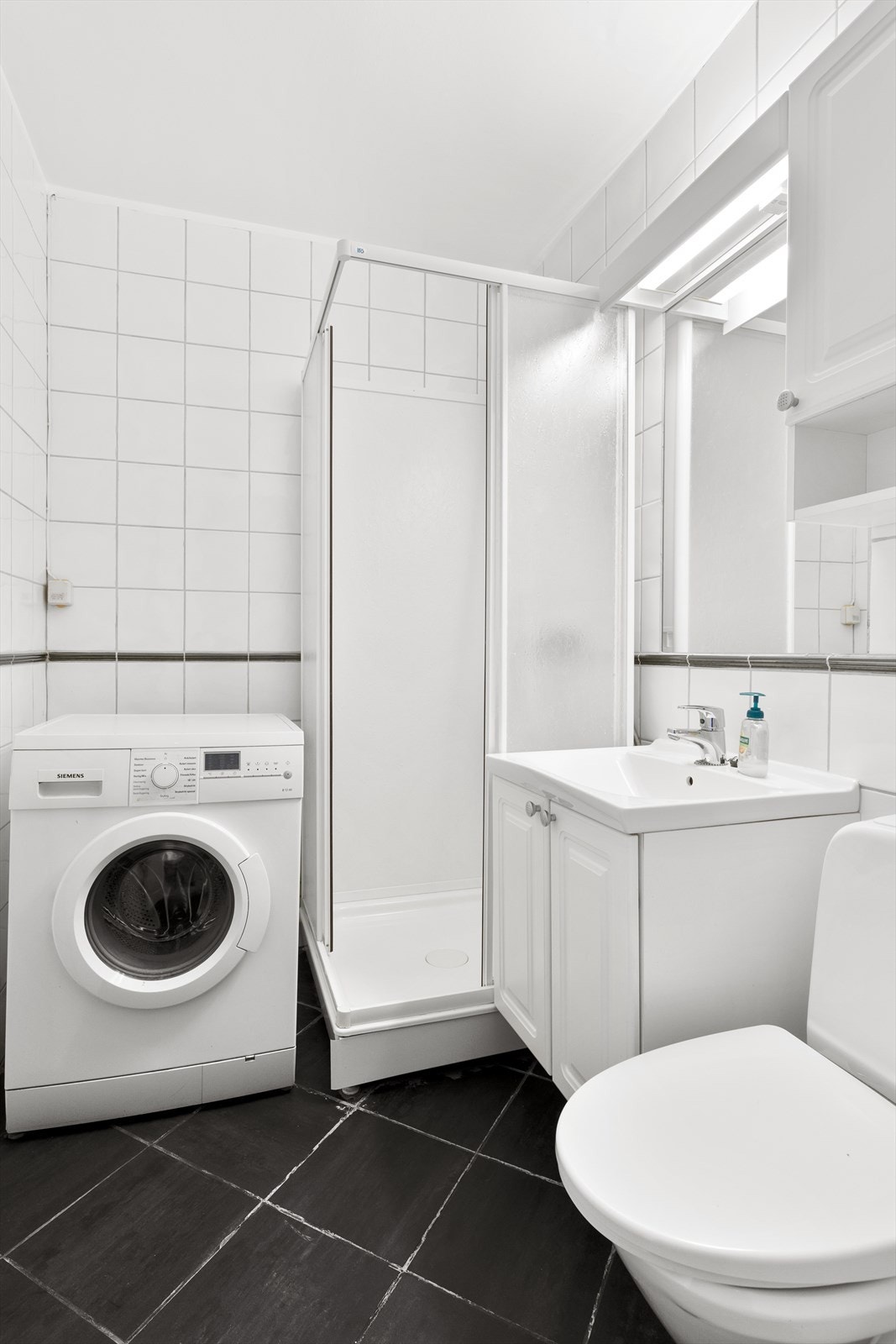 Baderomsinnredning med servant på underskap og speil med skap og belysning over. Installasjoner med dusjkabinett, toalett og opplegg for vaskemaskin. Galleribilde