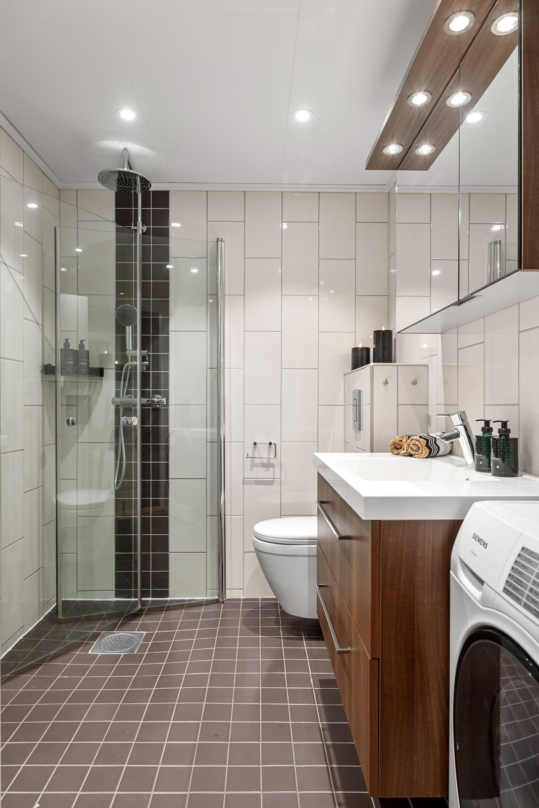 Her er det grå gulvfliser over gulvvarme og beige fliser på veggene med en sort, flislagt stripe i dusjhjørnet. Galleribilde