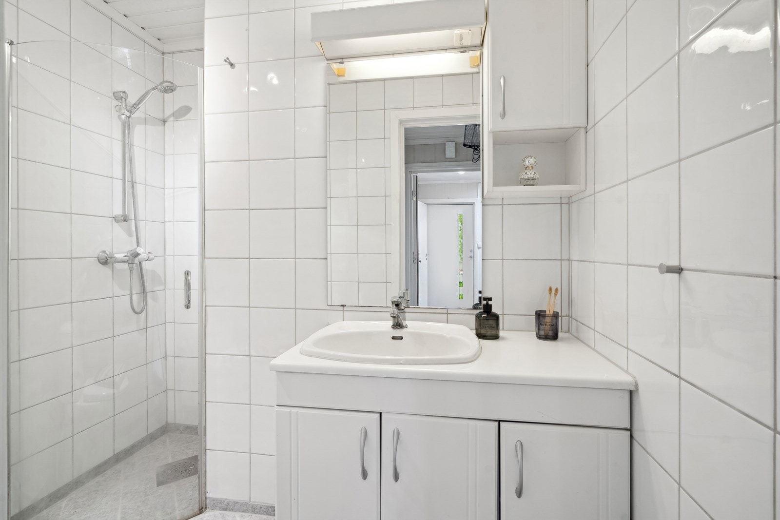 Flislagt baderom med lys innredning med nedfelt servant i heldekkende plate. Det er montert et toalett og dusjvegger. Galleribilde