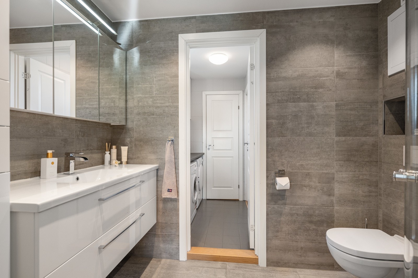 Badet har moderne farger og materialer. Det er innredet med dusjhjørne, vegghengt toalett og helstøpt servant med skuffer under. Galleribilde