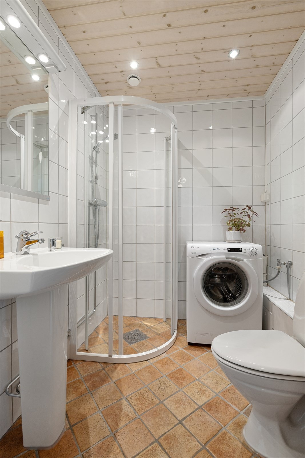 Badet har gulvstående servant med speil og belysning over, gulvstående toalett, dusjhjørne og opplegg for vaskemaskin. Galleribilde