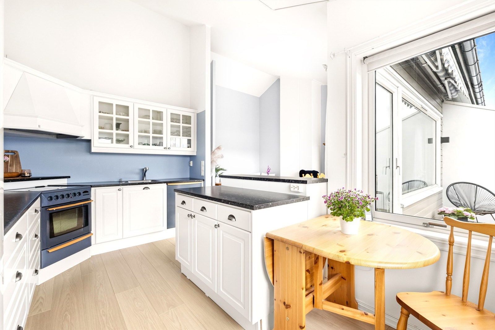 Innredningen er bygget i en klassisk L-form med hvite profilerte fronter, soft-close skuffer og skap, og benkeplater i sort og grått laminat. Det
er også plass til et bord for to i vindushjørnet. Galleribilde