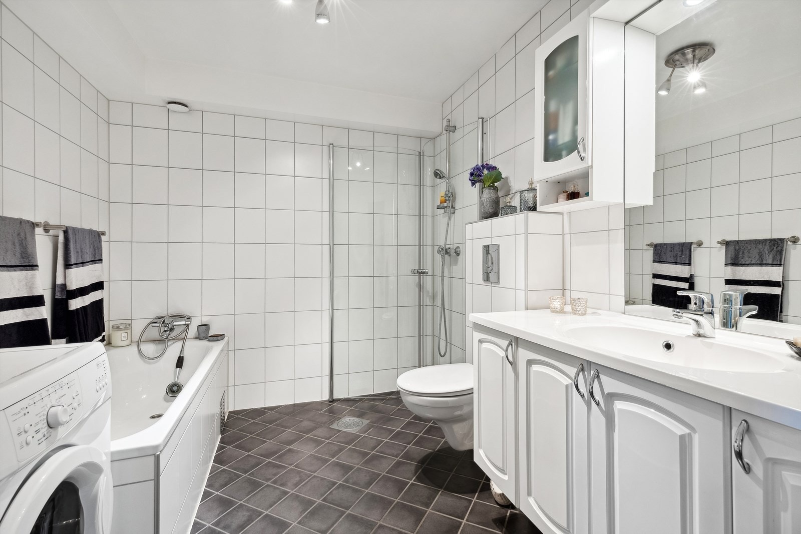 Badet er utstyrt med varmekabler, badekar, dusjhjørne med innfellbare glassvegger, vegghengt toalett og servant med tilhørende skap og speil. Galleribilde