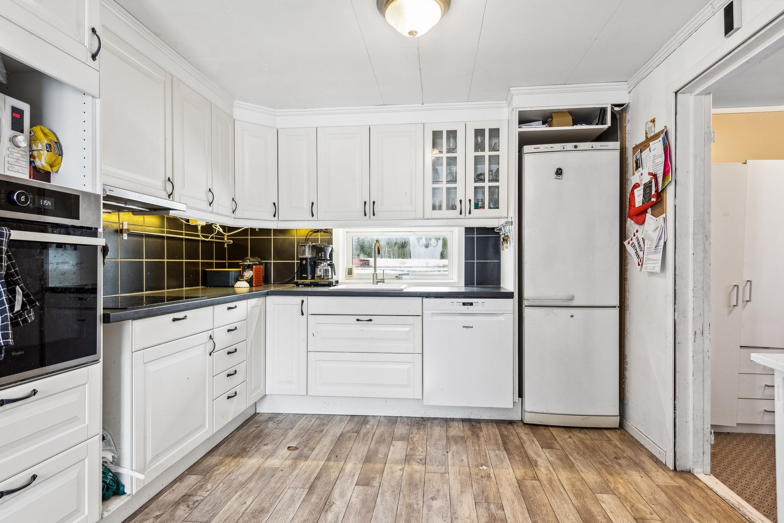 Kjøkken med lysmalte vegger og laminat på gulvet. Kjøkkeninnredning i L-formasjon med lyse profilerte fronter og laminerte benkeplater. Svarte fliser på veggflatene over kjøkkenbenkene. Galleribilde