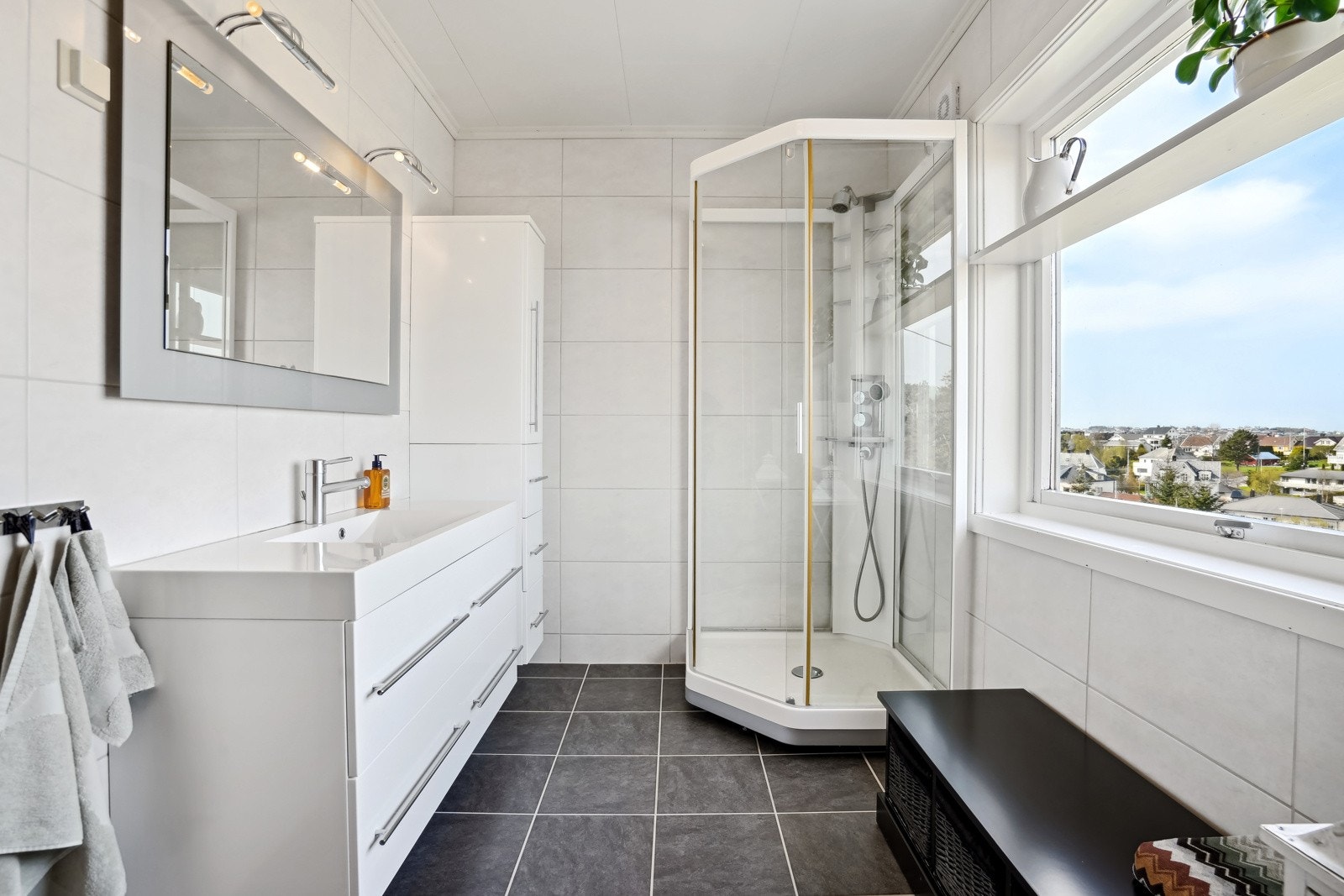 Lyst flislagt bad med elektriske varmekabler i gulv. Baderomsinnredningen har god plass til oppbevaring av velværeprodukter og håndklær. Galleribilde