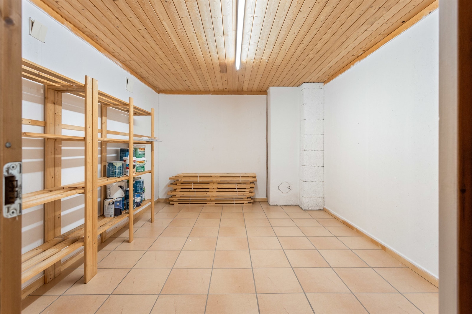 En bod i kjelleren har hvitmalte vegger og lyse fliser på gulvet. Bodrommet er innredet med hyller hvor du kan oppbevare ting. Galleribilde