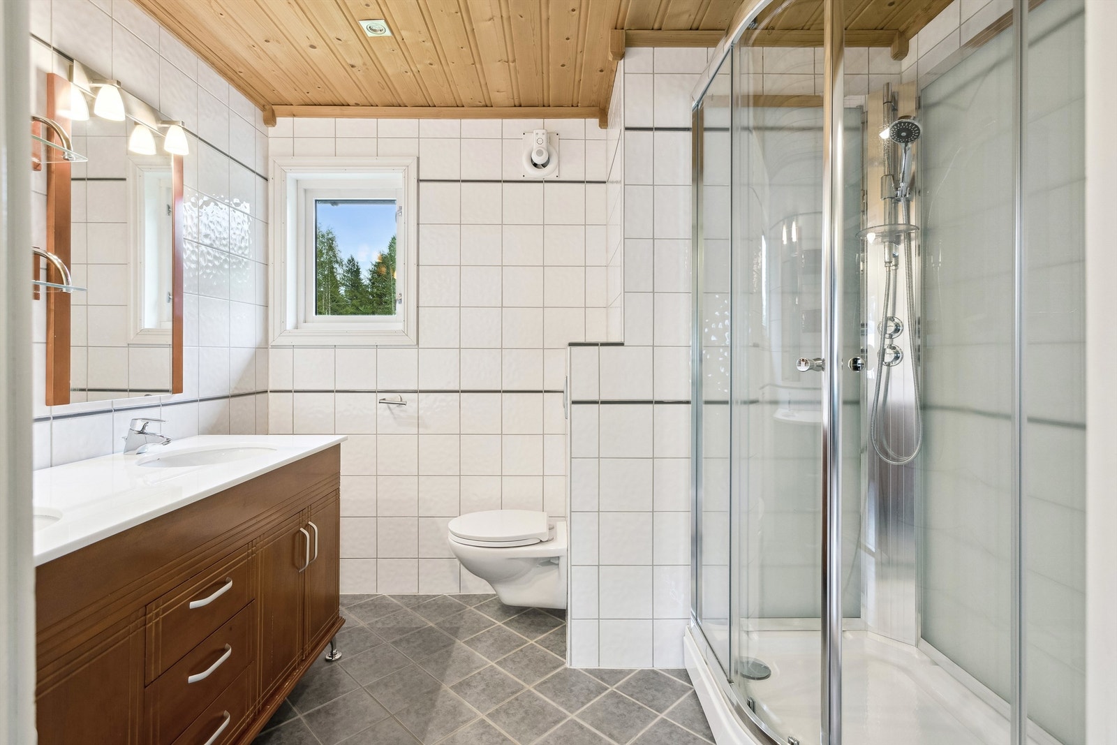 Her er det dobbelt servant i brun skap-/skuffeinnredning, vegghengt speil, dusjkabinett og veggmontert toalett. Galleribilde